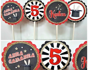 May include: Set of six black and red cupcake toppers with gold glitter accents. The toppers feature a magician's hat, stars, the number 5, and the words "Abra Cadabra".
