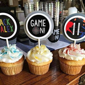 May include: Three cupcakes decorated with white frosting and colorful sprinkles. Each cupcake has a round paper topper with black and white text. The toppers read "Jack & Alex", "Game On!" and "Game On!"