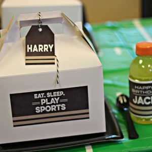 Puede incluir: Una caja de regalo blanca con una etiqueta en blanco y negro que dice "Eat. Sleep. Play. Sports." Una etiqueta con el nombre "Harry" est&aacute; unida a la caja. Un mantel estampado en verde y negro est&aacute; en el fondo.