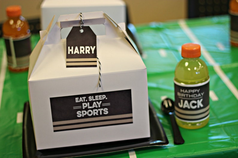 Puede incluir: Una caja de regalo blanca con una etiqueta en blanco y negro que dice "Eat. Sleep. Play. Sports." Una etiqueta con el nombre "Harry" est&aacute; unida a la caja. Un mantel estampado en verde y negro est&aacute; en el fondo.