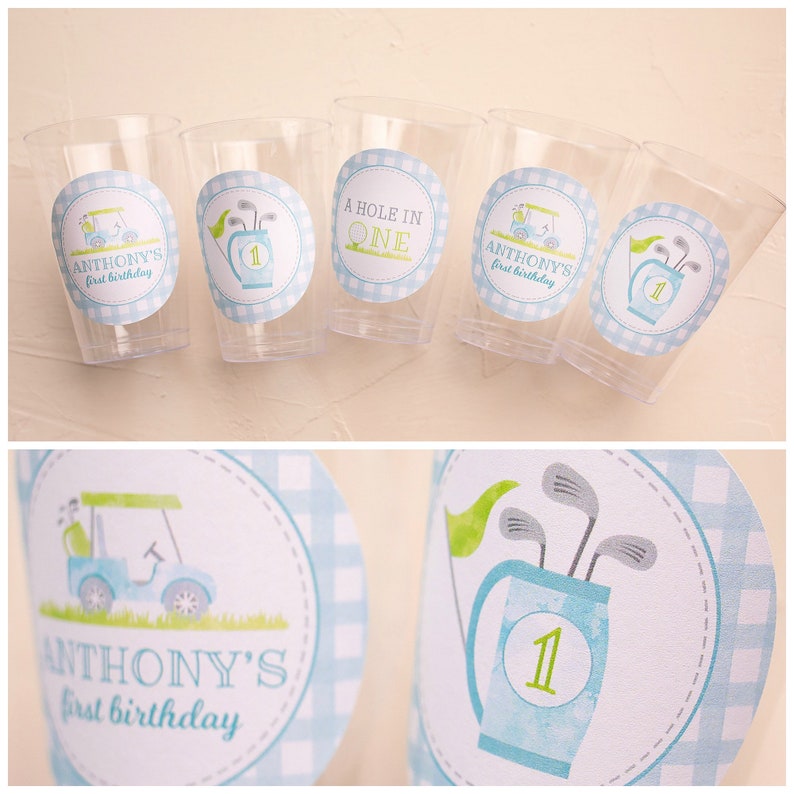 May include: Clear plastic cups decorated with golf-themed stickers. The stickers display a golf cart, golf clubs, and the text "ANTHONY'S first birthday" and "A HOLE IN ONE" in blue and green.
