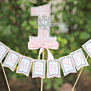 May include: A pink and gray "1st birthday" cake topper with a banner spelling "EMMERSON". The number one is decorated with snowflakes. The banner and number are attached to wooden sticks, perfect for a first birthday celebration.