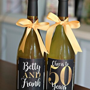 May include: Two wine bottles with black labels and gold lettering. One bottle says "Betty and Frank" and the other says "Cheers to 50 Years".