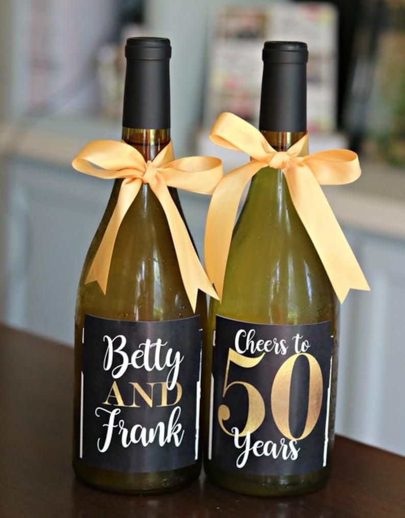 May include: Two wine bottles with black labels and gold lettering. One bottle says "Betty and Frank" and the other says "Cheers to 50 Years".