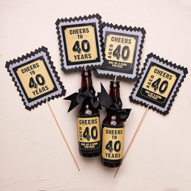May include: Two brown glass beer bottles with black and gold labels that say "Cheers to 40 Years" and "Cheers Aged 40 One of a Kind Vintage". The bottles are decorated with black ribbon and are on a white background. There are four black and gold party decorations with the same text as the bottles.