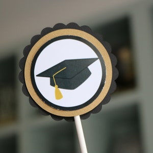 May include: Black and gold graduation cap cupcake topper. The topper features a black graduation cap with a gold tassel on a white circle with a gold and black border.