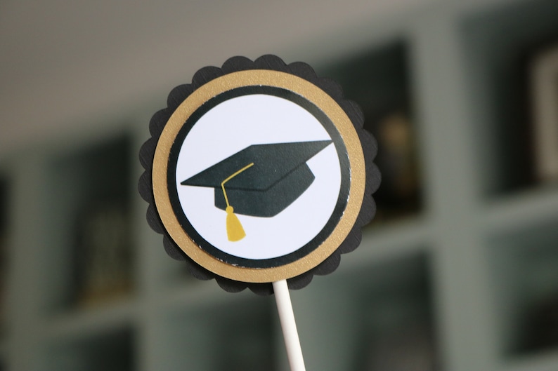 May include: Black and gold graduation cap cupcake topper. The topper features a black graduation cap with a gold tassel on a white circle with a gold and black border.