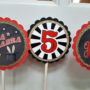 May include: Three red, black, and gold cupcake toppers with scalloped edges. The toppers feature a black background with gold glitter. One topper says "ABRA CADABRA" with a black top hat and white rabbit ears. Another topper has a black and white striped background with a red number "5". The last topper says "Kaden" with a black and white magic wand.