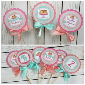 May include: Set of 8 pink and teal round paper cupcake toppers with scalloped edges. The toppers feature a pancake breakfast theme with illustrations of pancakes, syrup, and pajamas. Some toppers have the text "Happy Birthday" and a name, such as "Victoria" or "Natalie". Other toppers have the text "Pancakes and Pajamas".