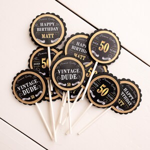 May include: Black and gold cupcake toppers with the text "Happy Birthday Matt", "Vintage Dude", and "Aged 50 Years".