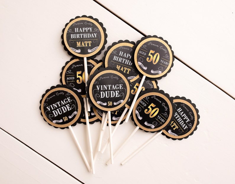 May include: Black and gold cupcake toppers with the text "Happy Birthday Matt", "Vintage Dude", and "Aged 50 Years".