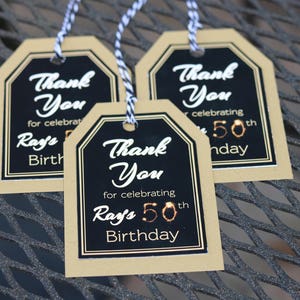 May include: Three gold and black thank you tags with black text that reads "Thank You for celebrating Ray's 50th Birthday".