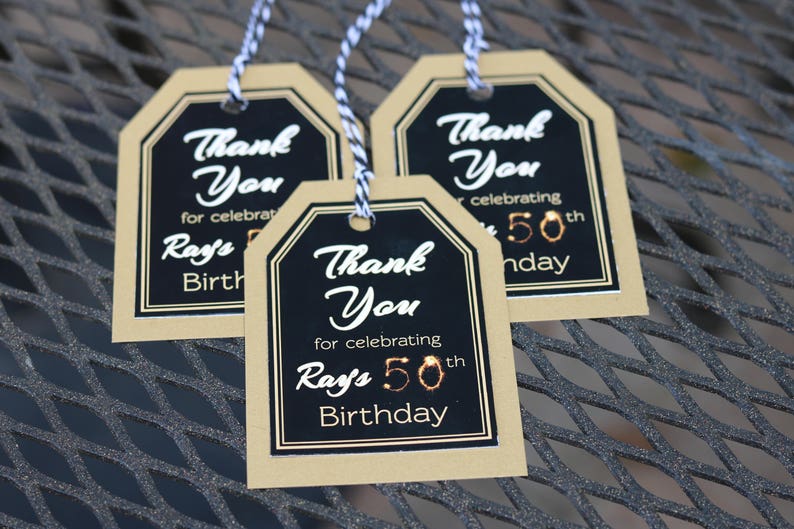 May include: Three gold and black thank you tags with black text that reads "Thank You for celebrating Ray's 50th Birthday".
