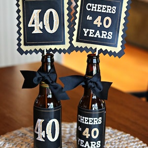 May include: Two small beer bottles decorated with black and gold labels. The labels say "Cheers to 40 Years". One bottle has a black bow tied around it. The bottles are sitting on a woven placemat.
