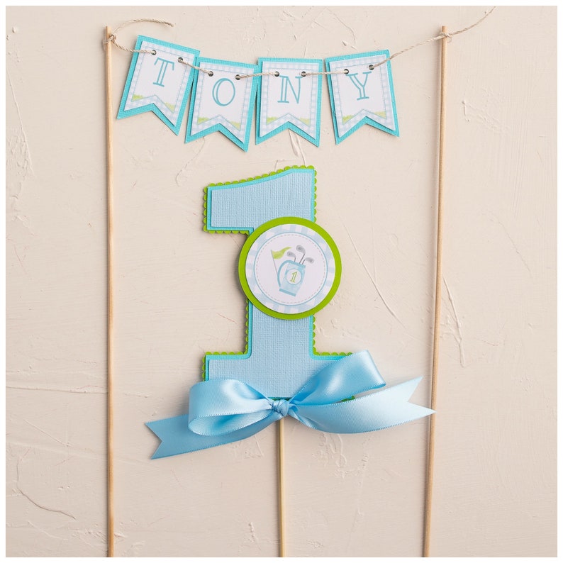 May include: A blue and green cake topper with the number one and a golf bag design. The topper also includes a banner that says "TONY".