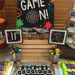 May include: A party table set up for a birthday party with a black and white sign that says "Game On!" with colorful paint splatters. There are two buckets filled with colorful gumballs, a wooden crate, and a tray of small clear tubes filled with colorful gumballs. The tubes have black tags that say "Team" on them. There are also boxes of "Power Pellets" on the table.