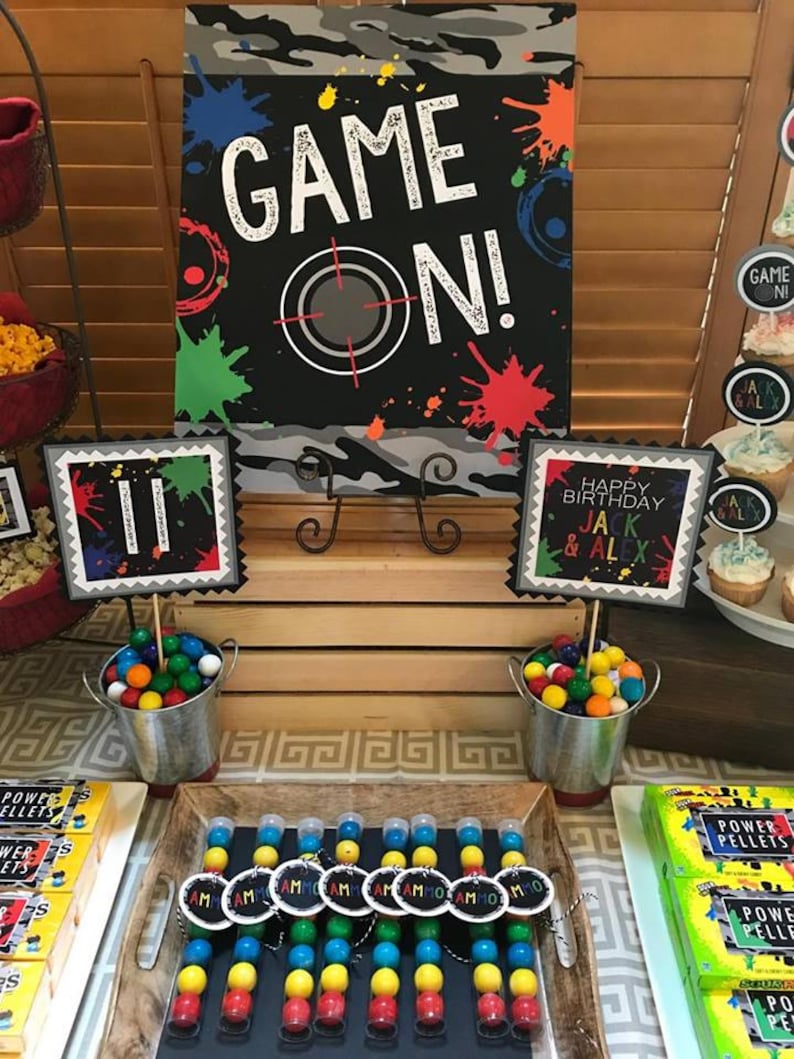 May include: A party table set up for a birthday party with a black and white sign that says "Game On!" with colorful paint splatters. There are two buckets filled with colorful gumballs, a wooden crate, and a tray of small clear tubes filled with colorful gumballs. The tubes have black tags that say "Team" on them. There are also boxes of "Power Pellets" on the table.