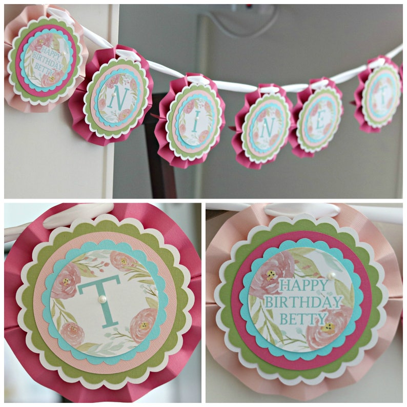 May include: A pink, green, and blue paper banner with the words "HAPPY BIRTHDAY BETTY" and individual letters spelling "ONE". Each circular element features floral designs and scalloped edges, strung on a white ribbon.
