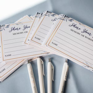 May include: A collection of white cards with gold borders and the text "Share Your Favorite Memory or Words of Love" in elegant script. The cards are decorated with blue floral accents and are accompanied by white pens.