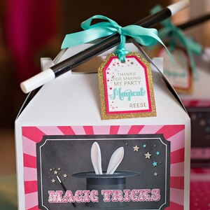 May include: White favor box with a pink and red striped design. The box has a black and white illustration of a magic hat with bunny ears and the text "Magic Tricks". A black and white magic wand is tied to the box with a turquoise ribbon and a gold glitter tag that says "Thanks for making my party Magical Reese".