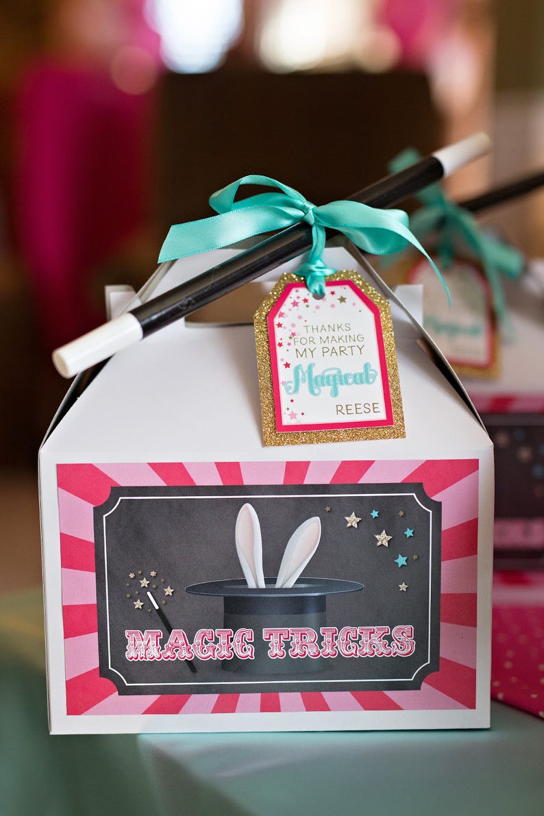 May include: White favor box with a pink and red striped design. The box has a black and white illustration of a magic hat with bunny ears and the text "Magic Tricks". A black and white magic wand is tied to the box with a turquoise ribbon and a gold glitter tag that says "Thanks for making my party Magical Reese".