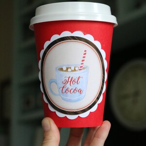May include: A bright red disposable paper cup with a white plastic lid. The cup features a circular label with a scalloped white border, displaying a watercolor illustration of a light blue mug filled with hot cocoa, marshmallows, and a red striped straw.