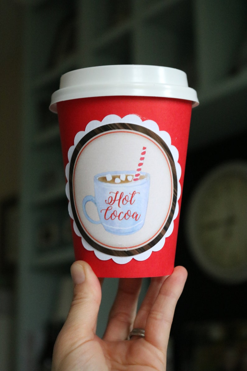 May include: A bright red disposable paper cup with a white plastic lid. The cup features a circular label with a scalloped white border, displaying a watercolor illustration of a light blue mug filled with hot cocoa, marshmallows, and a red striped straw.
