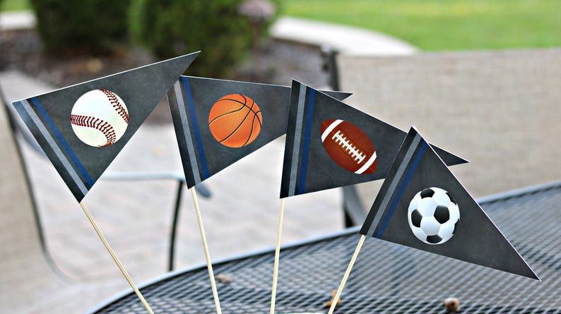 May include: Four gray pennant flags with blue stripes and different sports ball designs. The flags are on wooden sticks and are arranged in a fan shape. The sports balls are a baseball, basketball, football, and soccer ball.