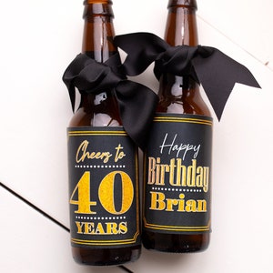 May include: Two brown glass beer bottles with black ribbon bows and personalized labels. The labels are black with gold lettering. One label says "Cheers to 40 Years" and the other says "Happy Birthday Brian".
