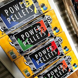 May include: Four boxes of M&M's candy stacked on top of each other. The boxes are yellow with a camouflage pattern and a black label that says "POWER PELLETS". The labels are different colors, including blue, green, red, and orange.