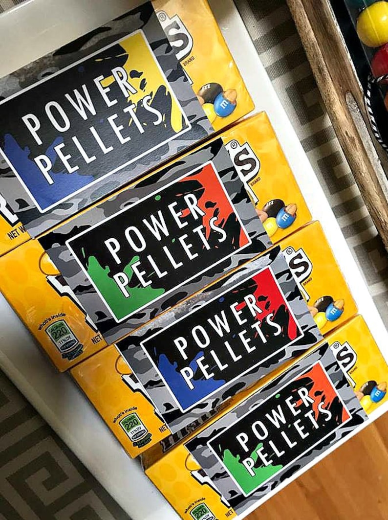 May include: Four boxes of M&M's candy stacked on top of each other. The boxes are yellow with a camouflage pattern and a black label that says "POWER PELLETS". The labels are different colors, including blue, green, red, and orange.
