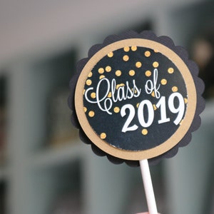 May include: Black and gold round cake topper with the text "Class of 2019" on a white stick.
