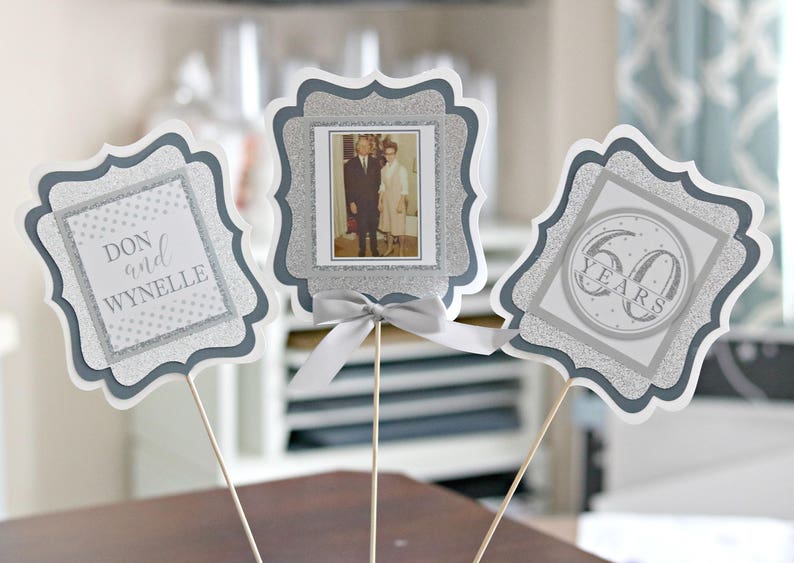 May include: Three silver glitter table centrepieces with a grey border. The centrepieces feature a photo of a couple, the names "Don and Wynnelie", and the text "60 Years".