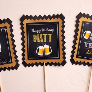 May include: Three black and gold party decorations with a scalloped edge. The center decoration says "Happy Birthday MATT" with two beer mugs. The left decoration says "50" and the right decoration says "50 YEARS LOVED".