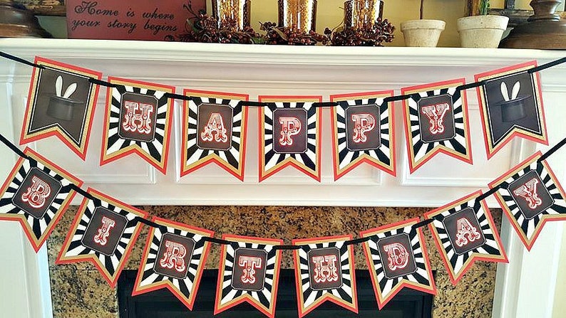 Op de afbeelding: Een zwart-wit gestreepte banner met rode accenten luidt "Happy Birthday". Elke letter staat in een rood kader met een witte achtergrond. De banner is versierd met zwarte hoge hoeden met witte konijnenoren.