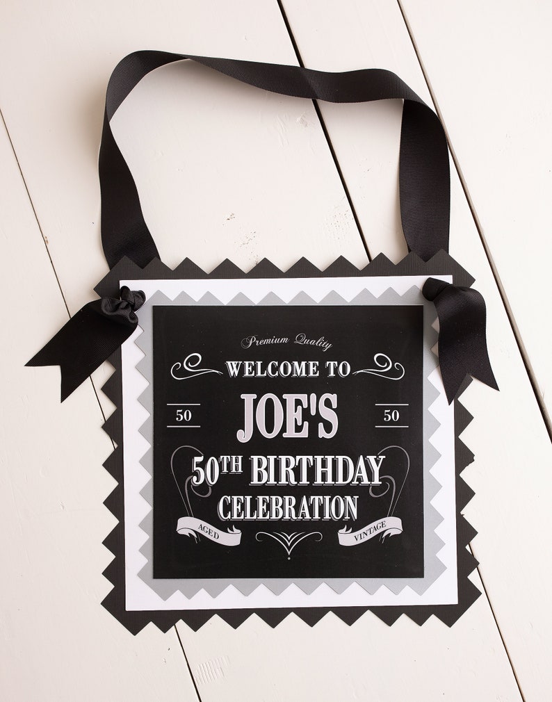 Puede incluir: Pancarta de cumplea&ntilde;os en blanco y negro con el texto "Welcome to Joe's 50th Birthday Celebration" y las palabras "Aged" y "Vintage" a cada lado del texto. La pancarta est&aacute; decorada con una cinta negra y tiene un borde festoneado.