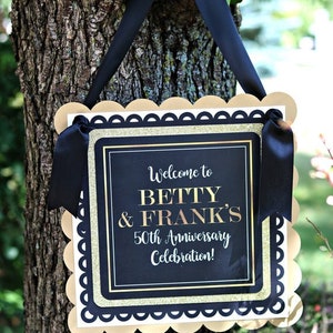 Puede incluir: Un cartel de bienvenida dorado y negro con bordes festoneados, colgado de una rama de &aacute;rbol. El cartel dice "&iexcl;Bienvenido a Betty & Frank's 50 aniversario de boda!"