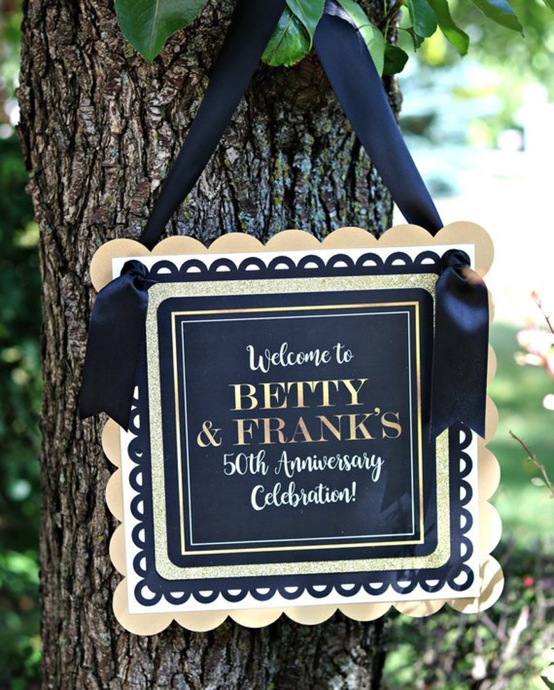 Puede incluir: Un cartel de bienvenida dorado y negro con bordes festoneados, colgado de una rama de &aacute;rbol. El cartel dice "&iexcl;Bienvenido a Betty & Frank's 50 aniversario de boda!"