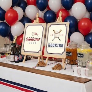May include: A baseball-themed party setup with red, white, and blue balloons in the background. Two signs on easels read "Oldtimes" and "Rookies." Beverages, baseball gloves, and cups are on a table.