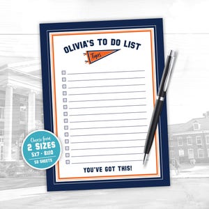 May include: A navy blue to-do list notepad with an orange and white border, and the text "OLIVIA'S TO DO LIST" at the top. A black pen rests on the notepad. The notepad includes the text "YOU'VE GOT THIS!"