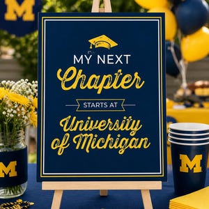 May include: A navy blue sign on a wooden easel with the text "My Next Chapter starts at University of Michigan" in yellow script. The sign is decorated with a graduation cap graphic. Yellow and navy blue balloons and party supplies are in the background.