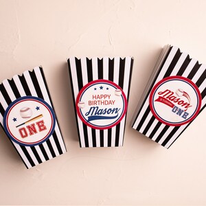 May include: Three black and white striped popcorn boxes with baseball-themed designs. The boxes have red, white, and blue accents. One box says "Happy Birthday Mason" with a baseball and bat graphic. Another box says "Mason is turning one" with a baseball graphic. The third box says "One" with a baseball and bat graphic.