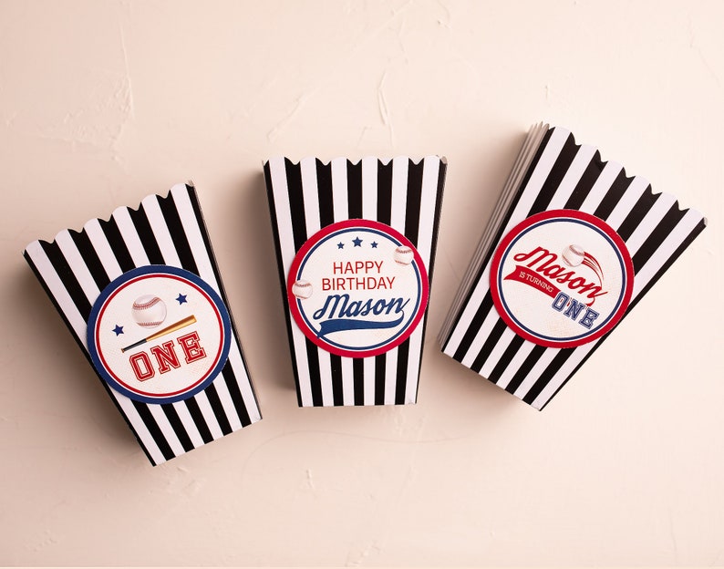 May include: Three black and white striped popcorn boxes with baseball-themed designs. The boxes have red, white, and blue accents. One box says "Happy Birthday Mason" with a baseball and bat graphic. Another box says "Mason is turning one" with a baseball graphic. The third box says "One" with a baseball and bat graphic.