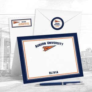 May include: Auburn University branded stationery set. Includes a folded card, envelope, address label, and a round sticker. The card and label feature the name "Olivia". The set has a navy blue and orange colour scheme.