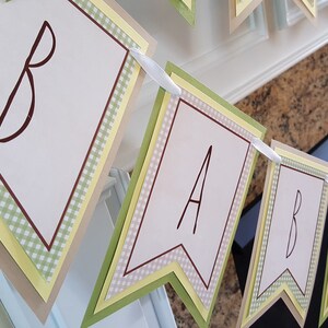 May include: A green, yellow, and brown banner with the word "BABY" spelled out in brown letters. The banner is made of paper and has a gingham pattern.