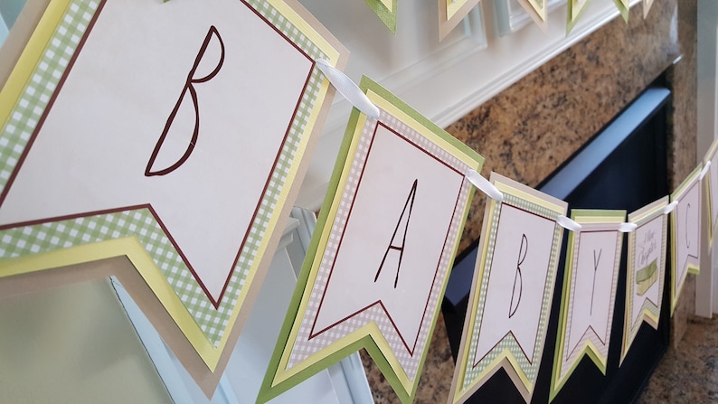 May include: A green, yellow, and brown banner with the word "BABY" spelled out in brown letters. The banner is made of paper and has a gingham pattern.