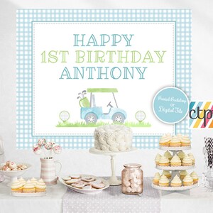 May include: A blue and green golf-themed first birthday party backdrop with the text "Happy 1st Birthday Anthony". The backdrop features a blue golf cart with a green golf bag and golf balls. The backdrop is printed on a blue and white gingham pattern.