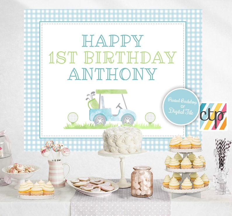 May include: A blue and green golf-themed first birthday party backdrop with the text "Happy 1st Birthday Anthony". The backdrop features a blue golf cart with a green golf bag and golf balls. The backdrop is printed on a blue and white gingham pattern.