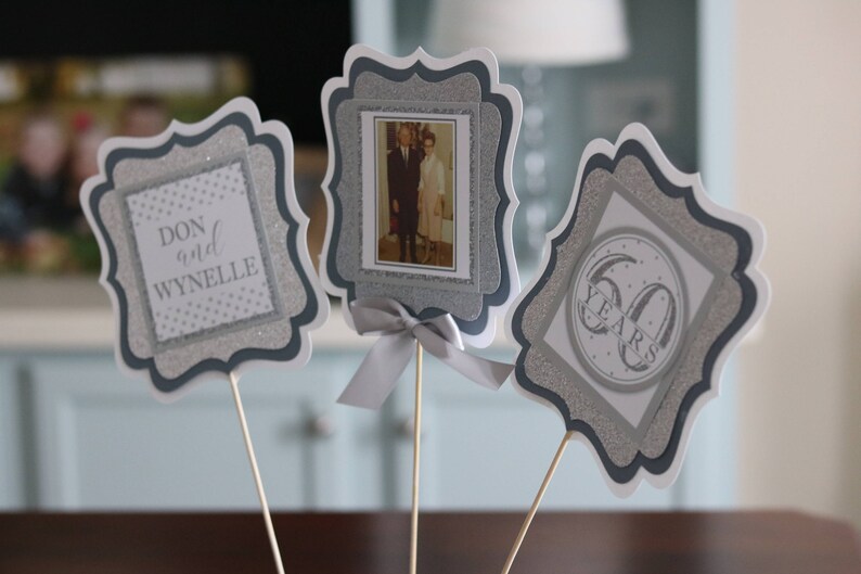 May include: Three silver and white table centrepieces with a decorative border. The centrepieces feature a photo of a couple, the names "Don and Wynnelie", and the text "60 Years".