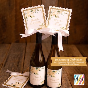 K&ouml;nnte beinhalten: Set aus drei goldenen und wei&szlig;en floralen Flaschenverschl&uuml;ssen mit dem Text "Cheers to 50 Years!" und "Olivia und Lucas". Die Verschl&uuml;sse sind mit einem wei&szlig;en Satinband verziert und bereit, auf Weinflaschen f&uuml;r eine 50. Jubil&auml;umsfeier platziert zu werden.